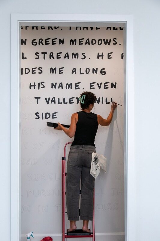 Brit Albin on a ladder painting scripture lettering onto the hallway wall